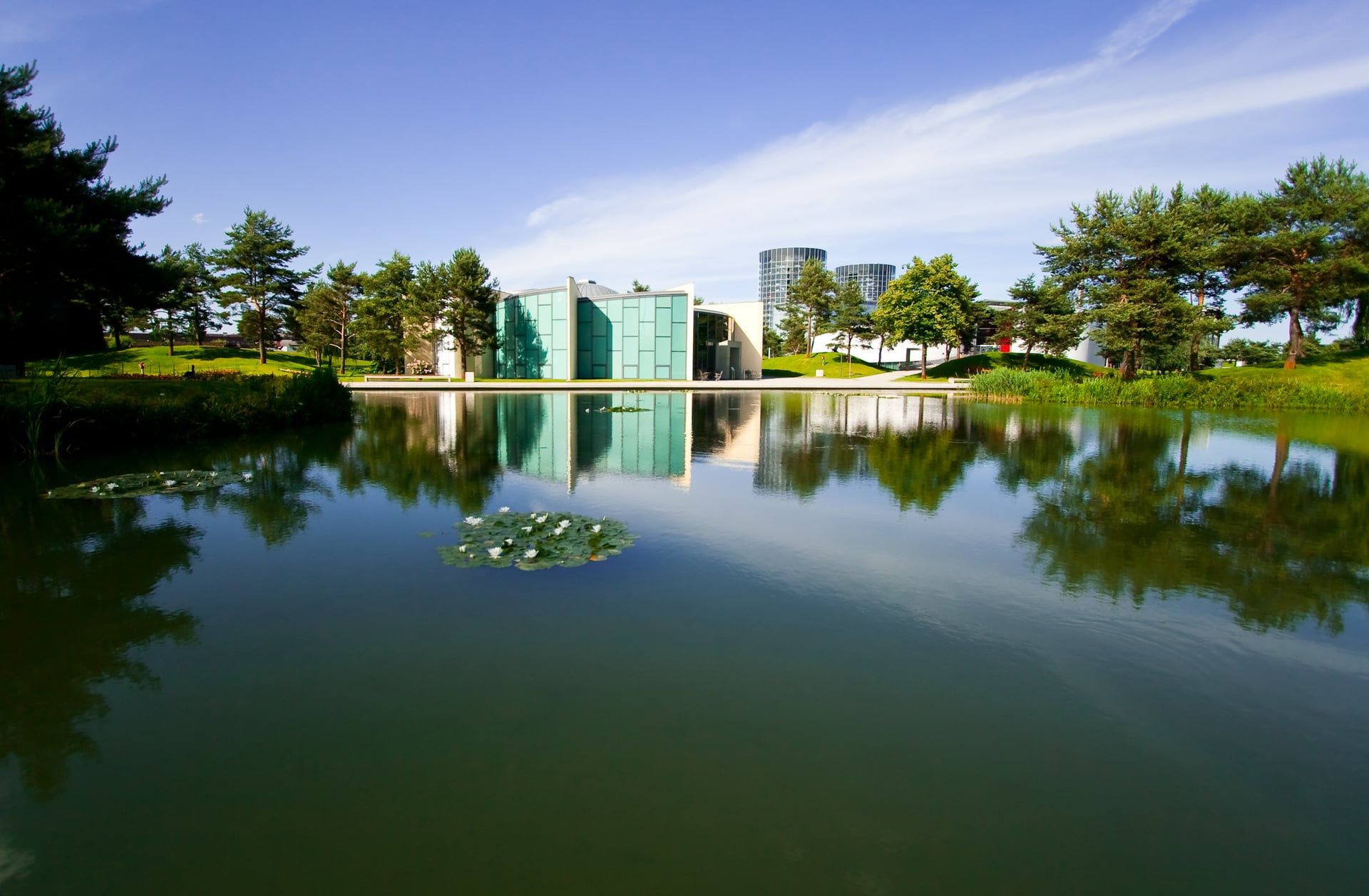 Modernes Museumsgebäude spiegelt sich in einem ruhigen Teich in einem japanischen Park mit Bäumen und blauem Himmel.