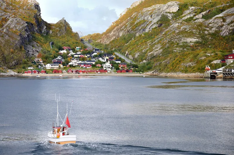 Fischerboot fährt durch einen norwegischen Fjord mit bunten Häusern an felsiger Küste im Hintergrund.