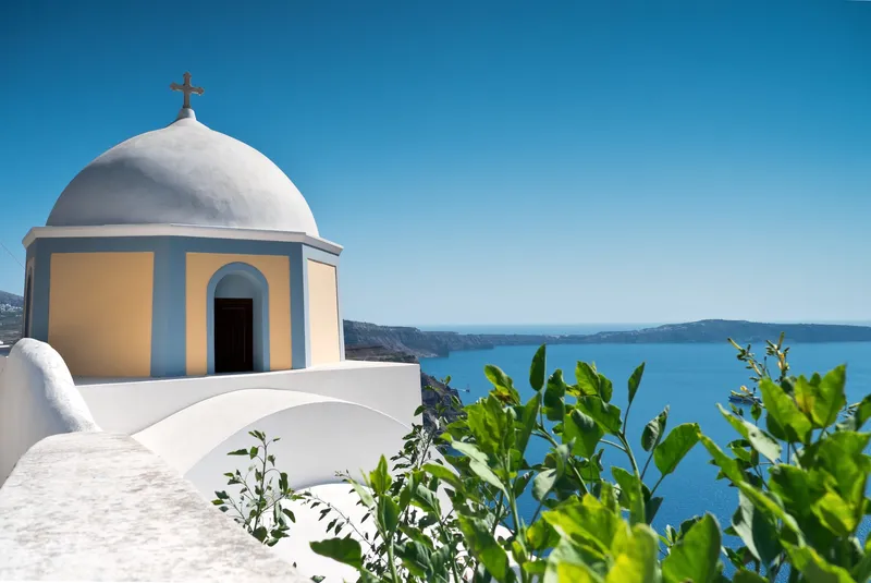 Weiße griechisch-orthodoxe Kirche mit Kuppelturm auf Santorin mit Blick auf die Caldera und das Meer.