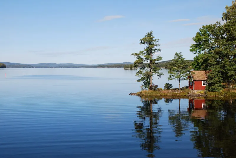 Kleines rotes Holzhaus an einem stillen skandinavischen See mit Spiegelung im Wasser und bewaldeten Hügeln.